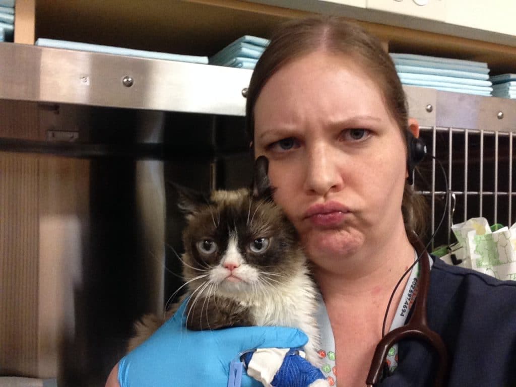 Photo of person named Michelle Crane in a vet tech uniform holding a grumpy cat and mimicking the cats facial expression.