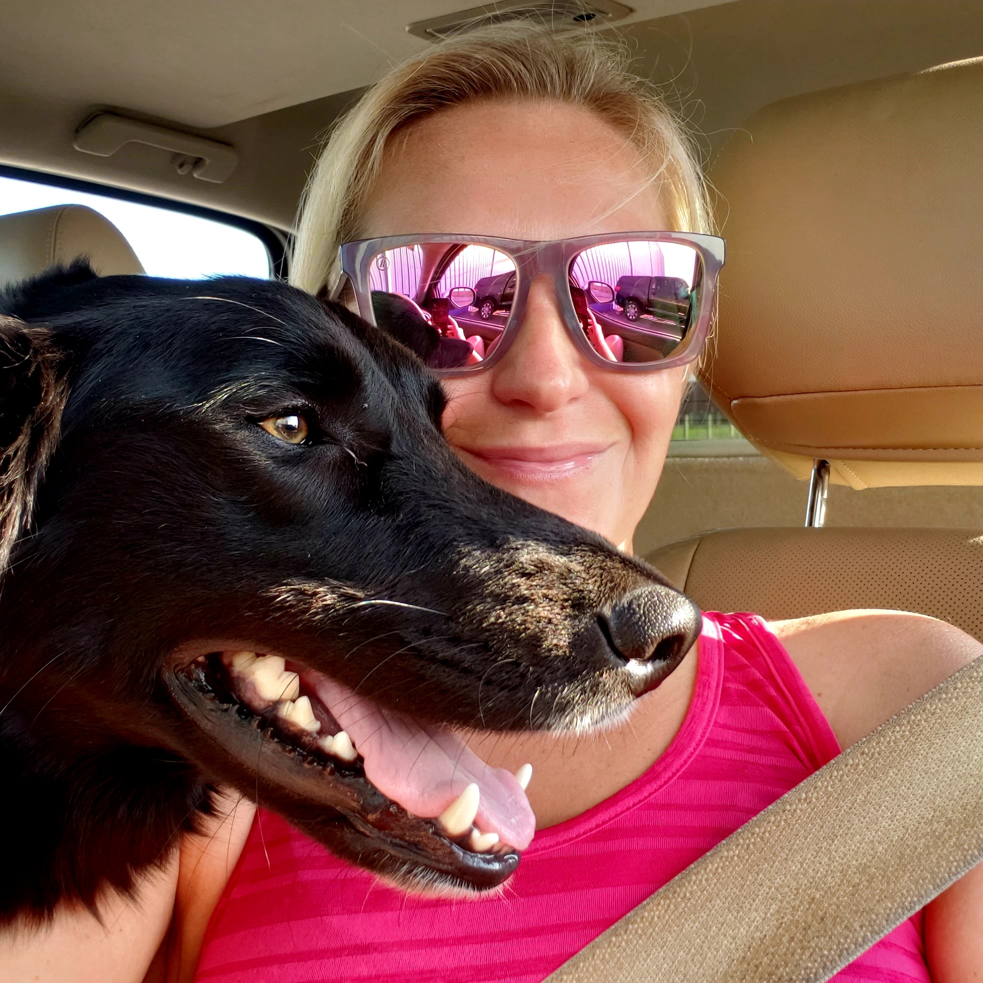 A blonde female veterinary professional wearing a bright pink tanktop pictured with the face of a large smiling black dog.