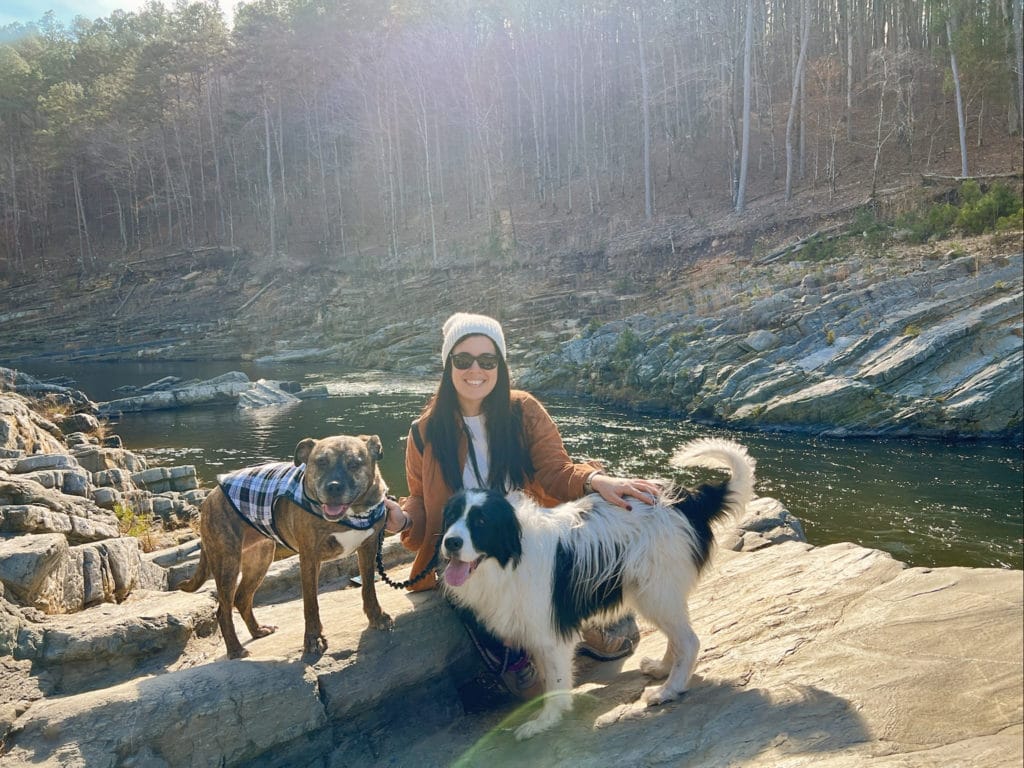 A woman with long brown hair hiking along a river with two dogs.