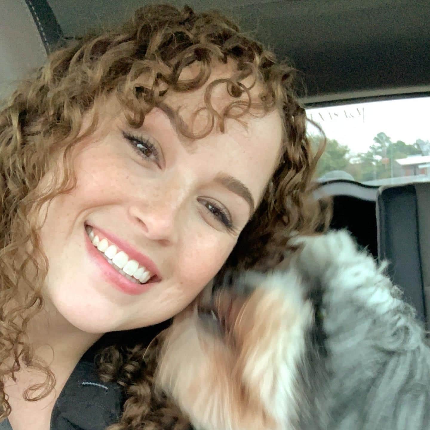 A young woman with curly brown hair receiving a kiss from a scruffy gray dog.