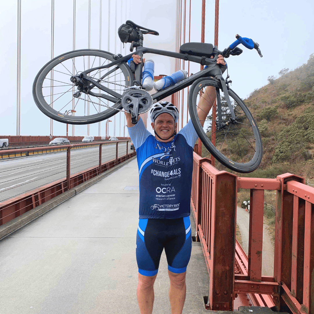 A man wears a helmet and a blue bicycling outfit that displays the NOMV logo on the front of the shirt. The man is holding up his road bicycle and smiling towards the camera. The Golden Gate Bridge is behind him.