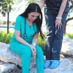 A women in turquoise scrubs looks down to the ground while sitting on a large rock. Another person in grey scrubs shows support to the woman by having their hand on her shoulder.